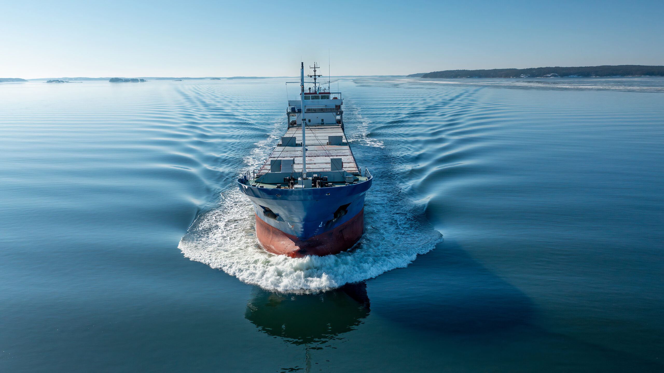 Aerial forward view of general cargo vessel making way ahead in Finnish archipelago during spring time. Glassy sea surface.